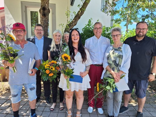 Ein Gruppenfoto von acht Personen im Freien. Sie halten Blumensträuße mit Sonnenblumen und Ordner. Eine junge Frau im weißen Kleid steht lächelnd im Zentrum. Die Szene deutet auf eine Verleihung oder feierliche Anerkennung hin.