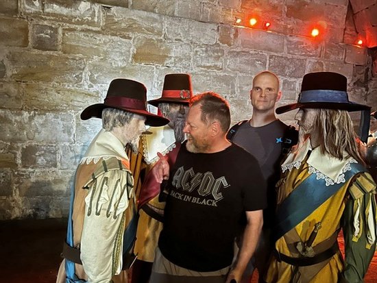 Ein Mann in einem schwarzen AC/DC-T-Shirt raucht eine Zigarette und steht zwischen zwei Personen in historischen Kostümen mit breiten Hüten. Ein weiterer Mann im Hintergrund blickt direkt in die Kamera vor einer Steinwand.