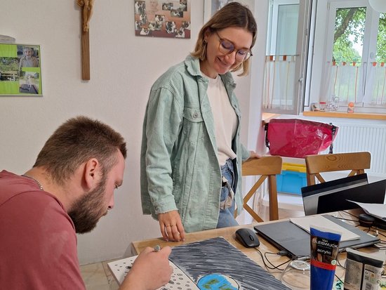 Ein bärtiger Mann malt an einem Tisch ein Bild mit einem blauen Planeten und einer Mondlandschaft. Eine Frau mit Brille steht lächelnd daneben und beobachtet ihn. Auf dem Tisch liegen Acrylfarben.