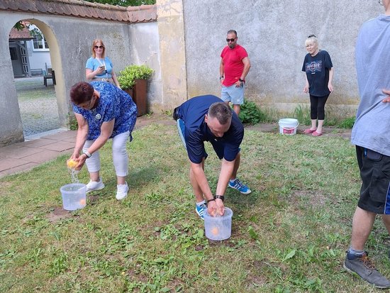 Zwei Personen beugen sich über Eimer auf einer Wiese und waschen sich die Hände. Im Hintergrund stehen mehrere Zuschauer und beobachten das Geschehen vor einem alten Gebäude.