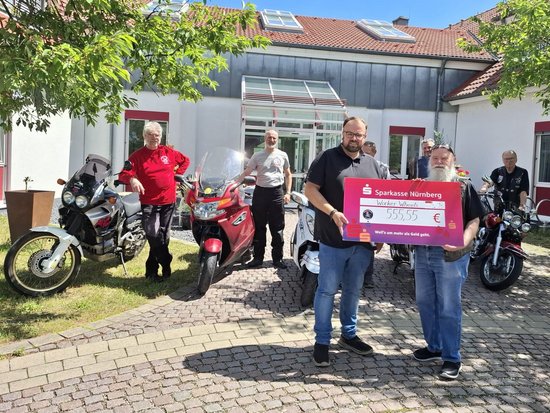 Zwei Männer halten einen großen Spendenscheck der Sparkasse Nürnberg über 555,55 Euro für „Worker Wheels". Im Hintergrund stehen mehrere Motorräder und weitere Personen vor einem Gebäude.