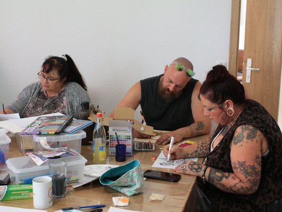 Drei Personen sitzen an einem Tisch und arbeiten konzentriert an künstlerischen Projekten. Vor ihnen liegen verschiedene Malutensilien wie Farben, Pinsel und Wasserflaschen. Es scheint sich um einen Kunstworkshop oder eine Malstunde zu handeln.